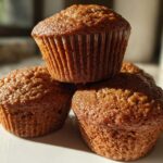 Close-up of three freshly baked, moist bran muffins stacked on a white plate, highlighted by sunlight.