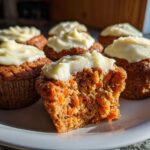 A close-up of several moist carrot cake muffins topped with cream cheese frosting, one is broken open showing shredded carrots.
