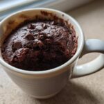 Close-up of a moist chocolate mug cake baked in a white mug, topped with chocolate chips.