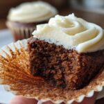Close-up of a moist gingerbread cupcake cut in half, showing the dark crumb and thick swirl of cream cheese frosting.