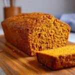 Close-up of a moist, freshly baked pumpkin bread loaf with one slice cut and resting beside it on a wooden board.