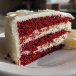 A thick slice of moist red velvet cake with three layers of deep red cake separated by white cream cheese frosting.