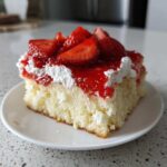A moist slice of jello poke cake topped with whipped cream, strawberry glaze, and fresh sliced strawberries.