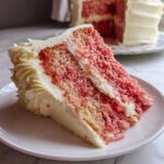 Close-up of a moist slice of strawberry cake with pink crumb and white frosting, served on a white plate.
