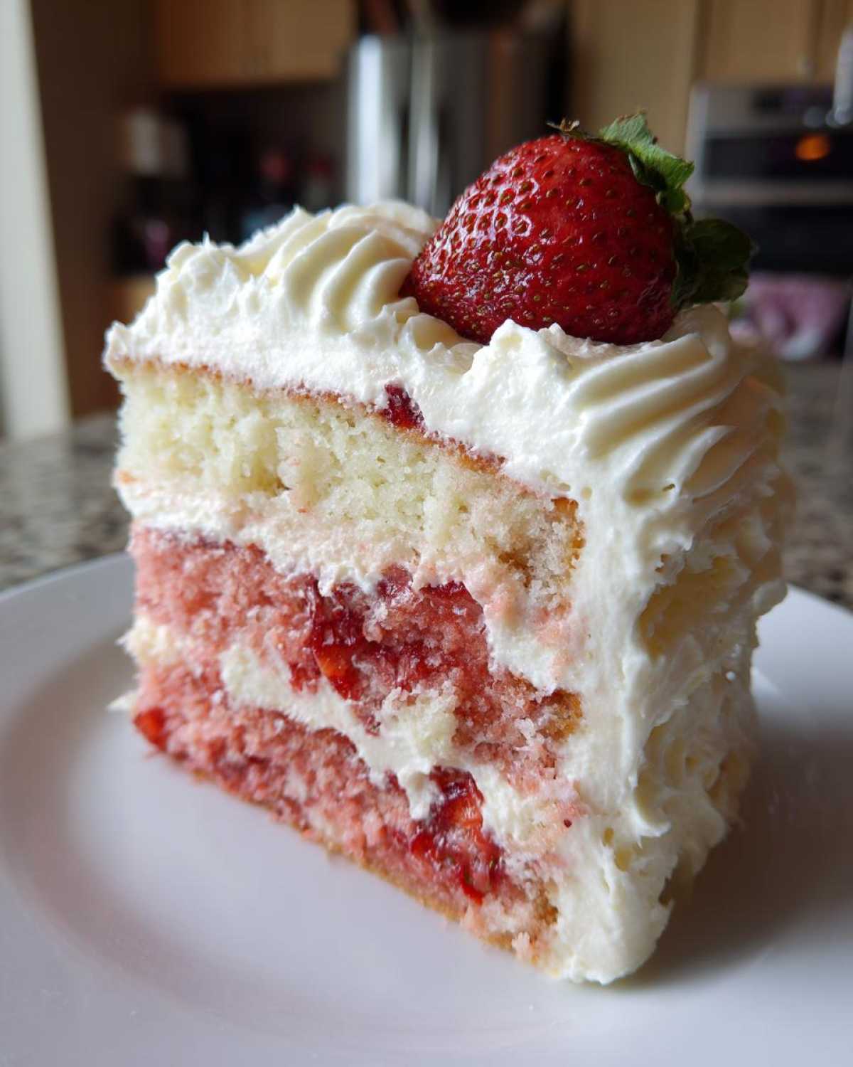 A tall slice of moist strawberry cake layered with cream filling and topped with whipped frosting and a fresh strawberry.