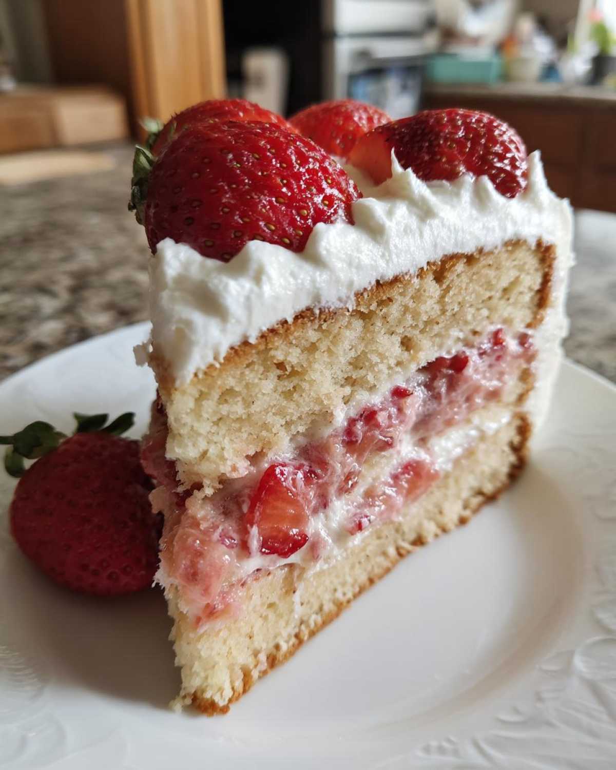 Close-up of a moist strawberry cake slice layered with cream and fresh strawberries on top.