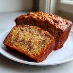 A moist slice of sugar free banana bread sits next to the remaining loaf, topped with pecans.