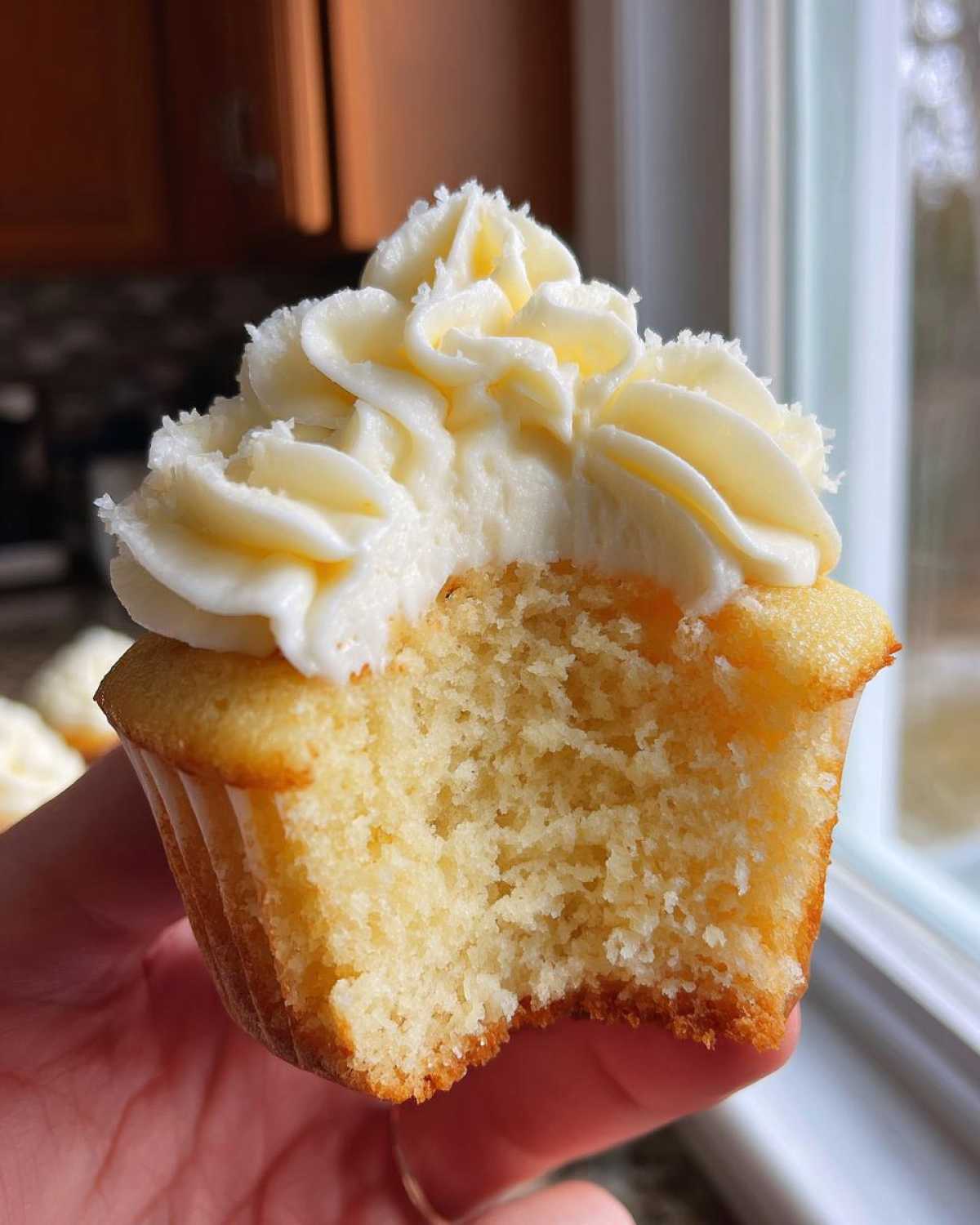 Close-up of a vanilla cupcake held in hand, showing a bite taken out to reveal the moist crumb and rich vanilla frosting.