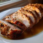 Slices of perfectly cooked pork loin roast resting in pan juices on a white platter, showing a golden, herb-crusted exterior.