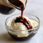 Close-up of rich chocolate syrup being poured from a glass pitcher onto scoops of vanilla ice cream.