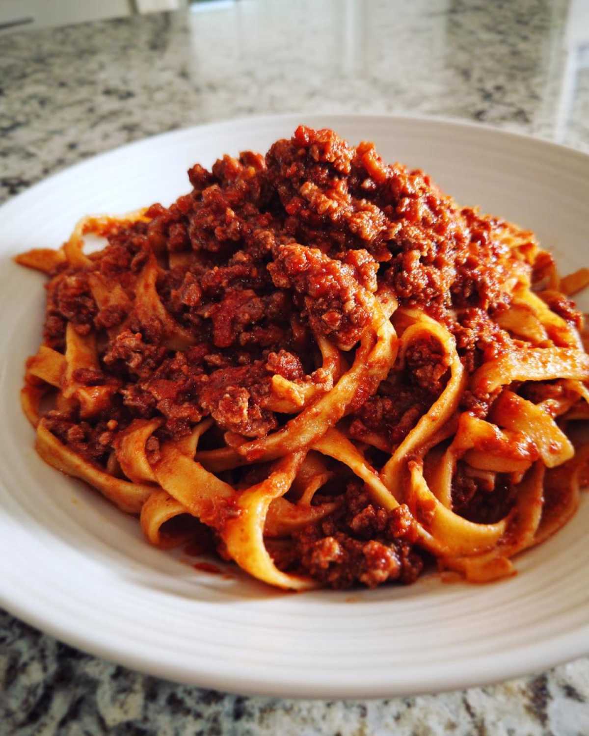 Close-up of thick, rich bolognese sauce generously covering a mound of fresh tagliatelle pasta in a white bowl.