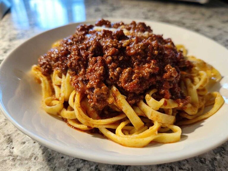 A close-up of fettuccine pasta generously topped with rich, dark red bolognese sauce.