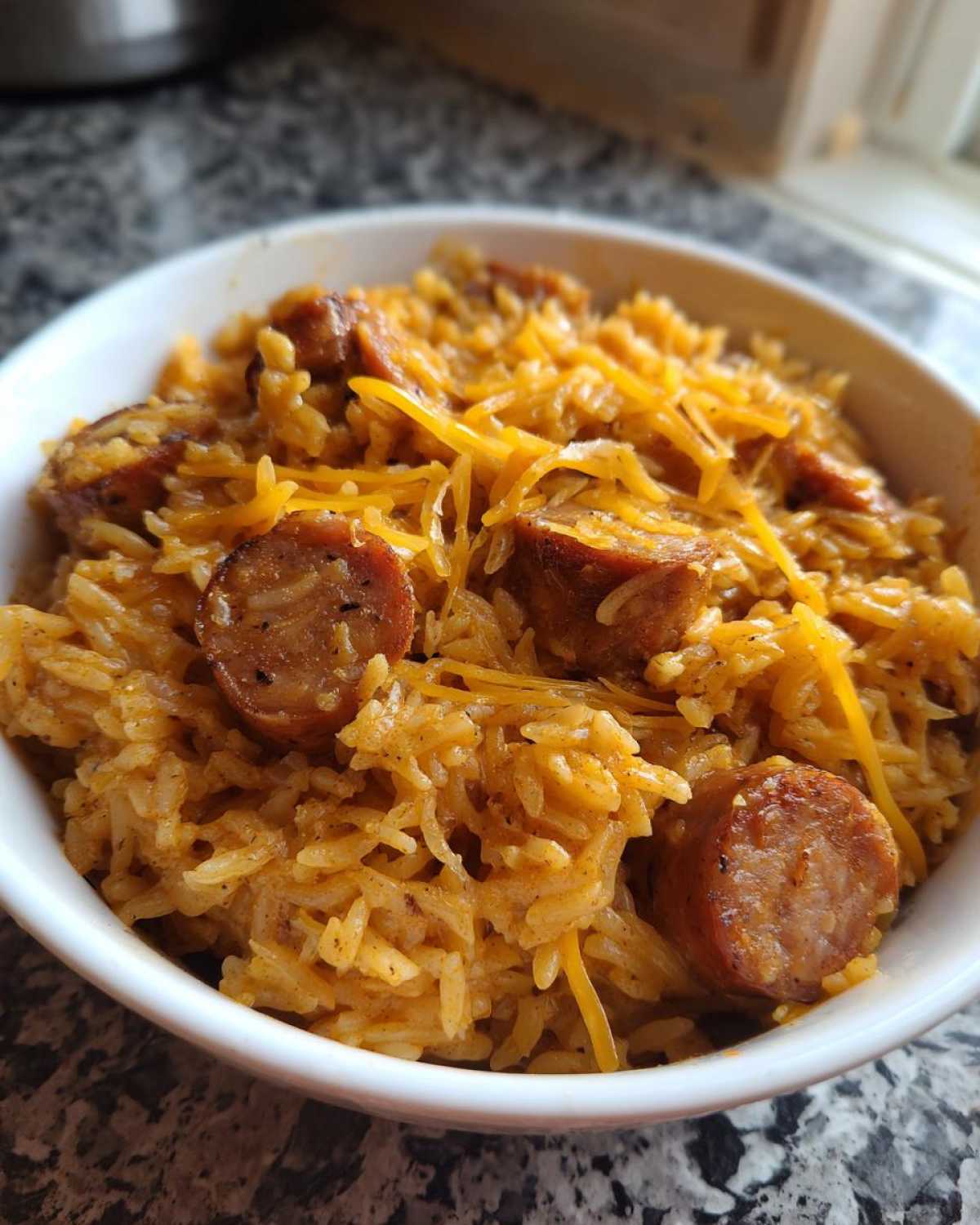Close-up of seasoned sausage and rice topped with shredded cheddar cheese in a white bowl.
