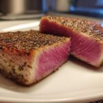 Two pieces of perfectly seared tuna steak, showing a vibrant pink rare center and a dark pepper crust, ready to eat.