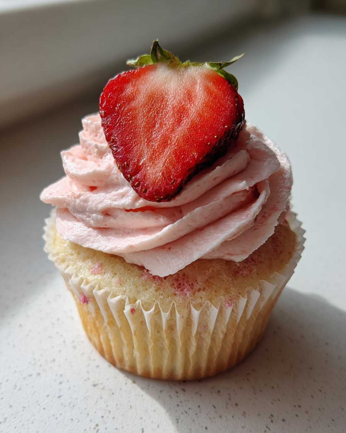 A close-up of one perfectly frosted strawberry cupcake topped with a fresh, halved strawberry.