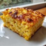 A close-up of a golden-brown slice of moist cornbread casserole, rich with corn kernels, served on a white plate.