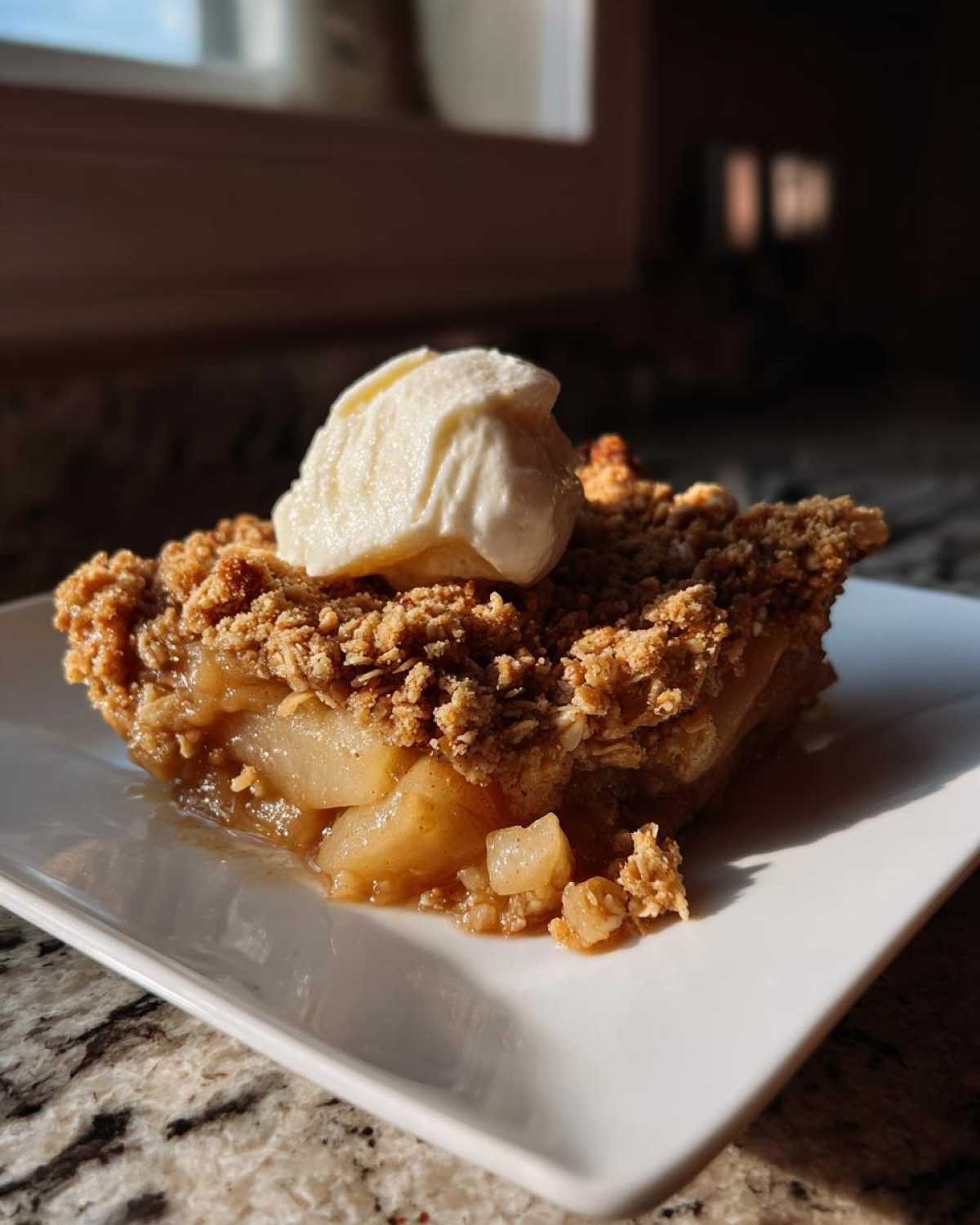 A warm slice of pear crisp with a generous oat topping and a scoop of vanilla ice cream on a white plate.