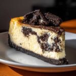 A rich slice of oreo cheesecake showing a marbled cream cheese filling, dark cookie crust, and topped with crushed Oreos.