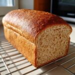 A golden-brown loaf of homemade whole wheat bread resting on a wire cooling rack.