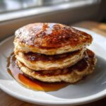 A stack of three fluffy sourdough discard pancakes drizzled generously with maple syrup on a white plate.