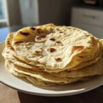 A stack of freshly made, slightly charred corn tortillas resting on a white plate in bright sunlight.