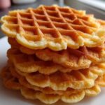 A tall stack of freshly made, golden brown pizzelle cookies with scalloped edges.