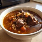 Close-up of a steaming white bowl filled with rich beef barley soup, showing chunks of beef, barley, and carrots.