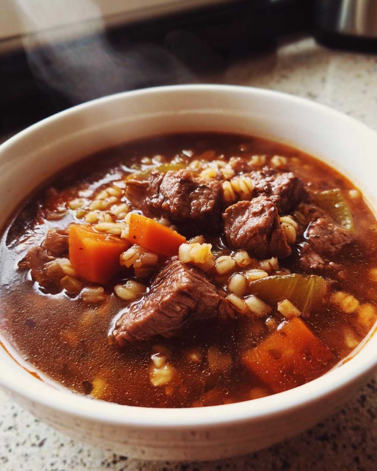 Close-up of a steaming white bowl filled with rich beef barley soup, showing chunks of beef, carrots, and barley.
