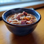 Close-up of a steaming blue bowl filled with rich Ham and bean soup, featuring white beans, diced ham, and carrots.