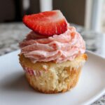 Close-up of a delicious strawberry cupcake with pink frosting and a fresh strawberry slice garnish; a bite is taken out.