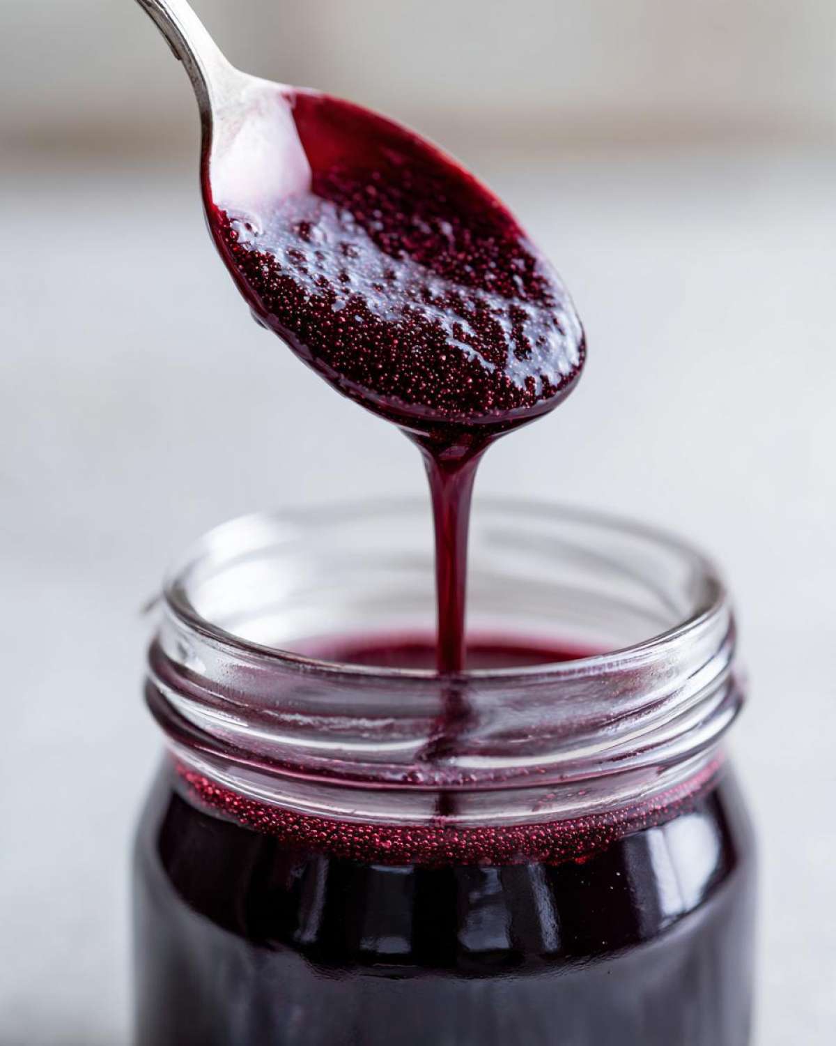 Close-up of thick, dark purple elderberry syrup recipe dripping from a spoon into a glass jar.