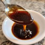 Close-up of thick, dark homemade teriyaki sauce dripping from a silver spoon into a small white bowl.
