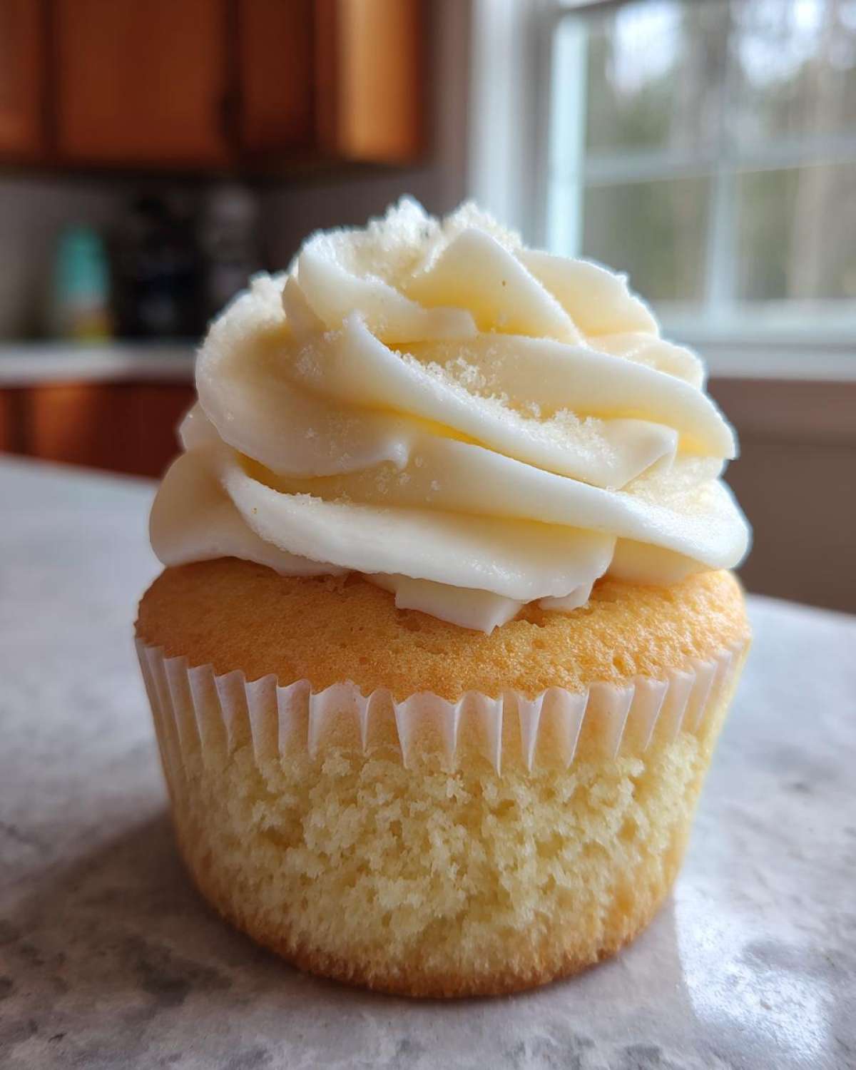 A single, moist vanilla cupcake topped with a generous swirl of white vanilla frosting and a sprinkle of sugar.