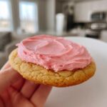 A hand holding a thick, chewy Copycat Crumbl Sugar Cookie topped with vibrant pink frosting.