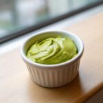 A small white ramekin filled with vibrant, smooth Creamy Avocado Cilantro Sauce, sitting on a wooden surface near a window.