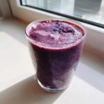 A close-up of a vibrant, thick Creamy Blueberry Banana Smoothie in a clear glass, sitting on a windowsill in bright sunlight.