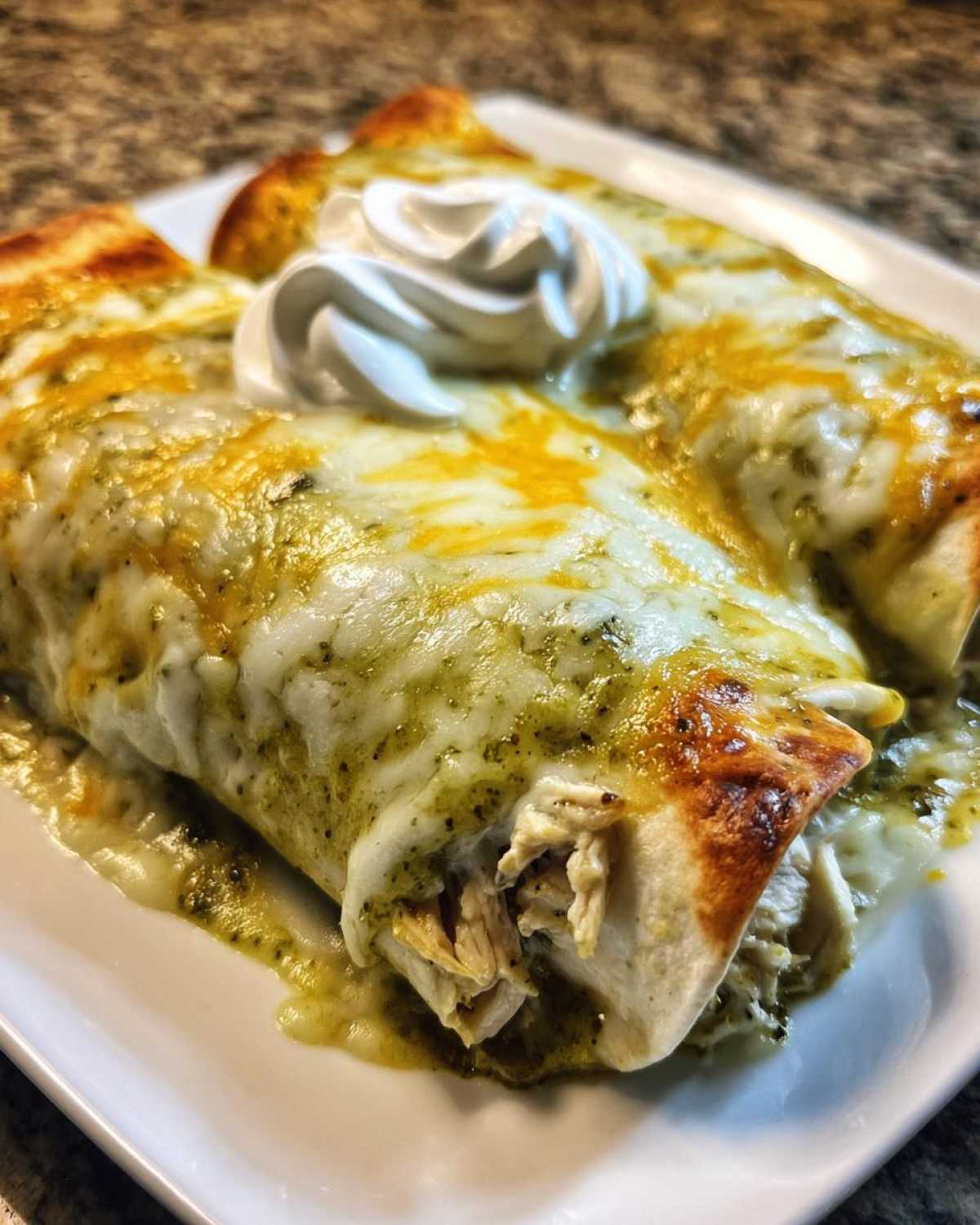 Close-up of two Creamy White Chicken Enchiladas smothered in green chile sauce and melted cheese, topped with sour cream.