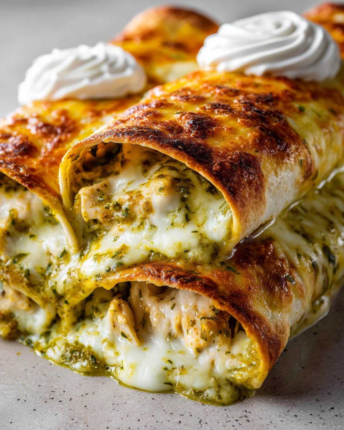 Close-up of baked Creamy White Chicken Enchiladas with Green Chile Sauce oozing melted cheese and topped with sour cream.