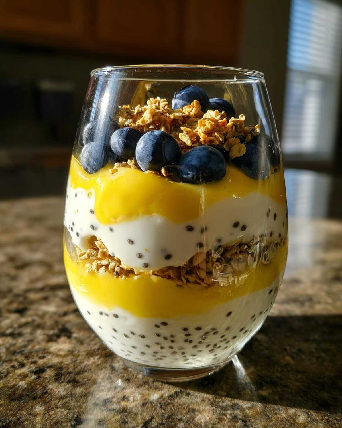 Close-up of an Easy Lemon Poppy Seed Yogurt Parfait layered with chia pudding, yellow lemon curd, granola, and topped with fresh blueberries.