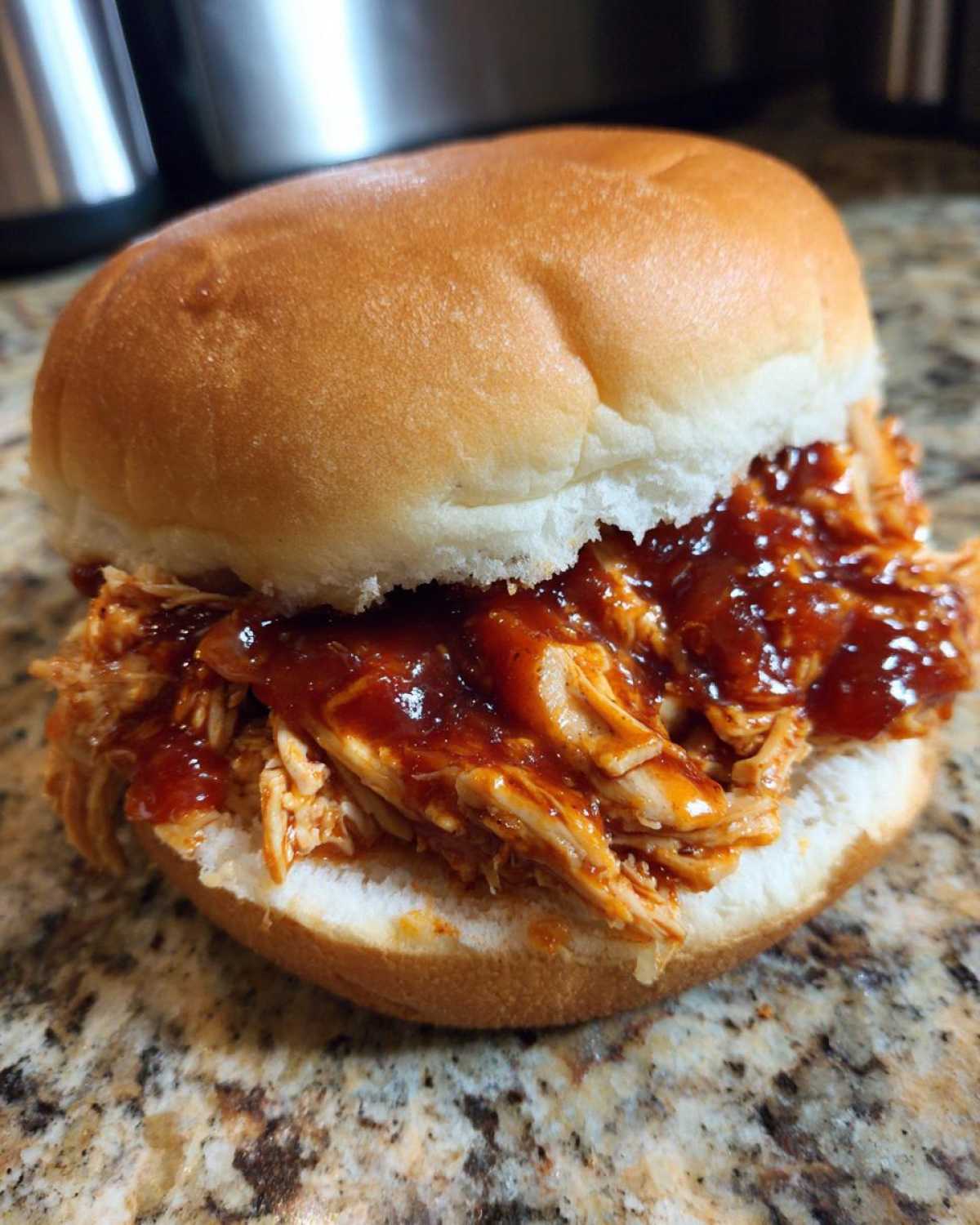 A close-up of a juicy Instant Pot BBQ Pulled Chicken Sandwich on a soft bun, sitting on a granite counter.