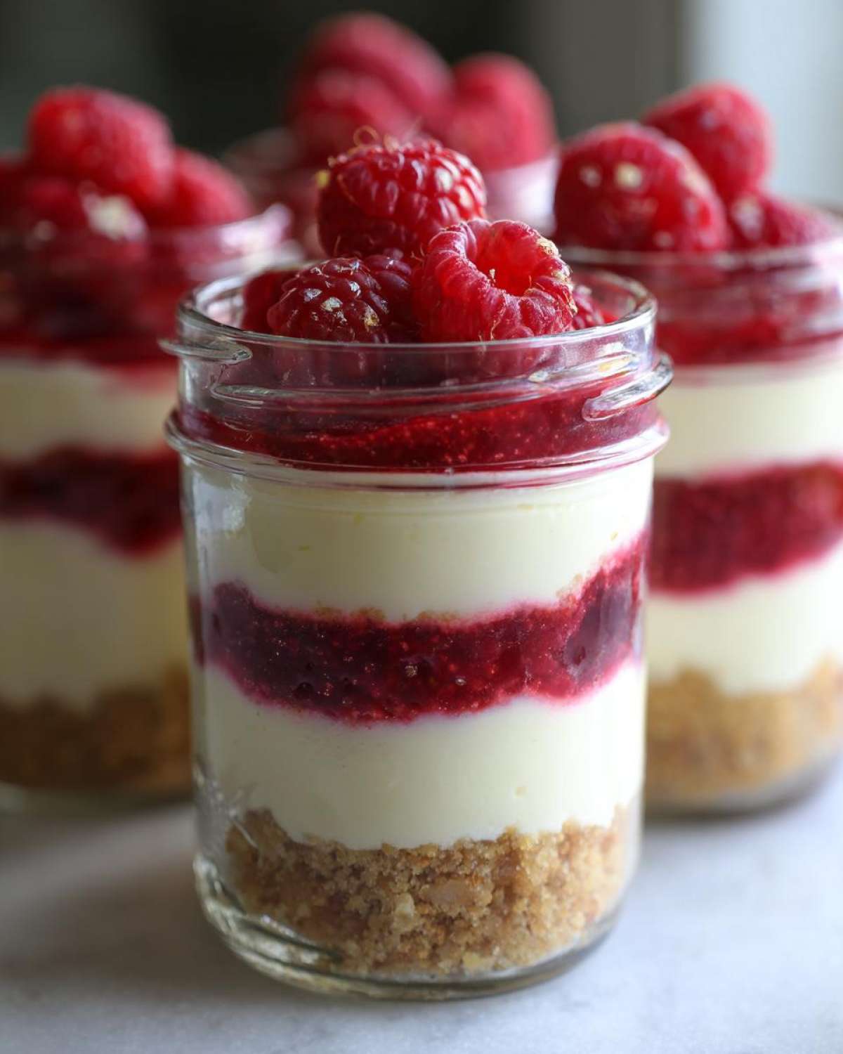 Close-up of No-Bake Raspberry White Chocolate Cheesecake Jars layered with crust, cream, and raspberry filling, topped with fresh raspberries.