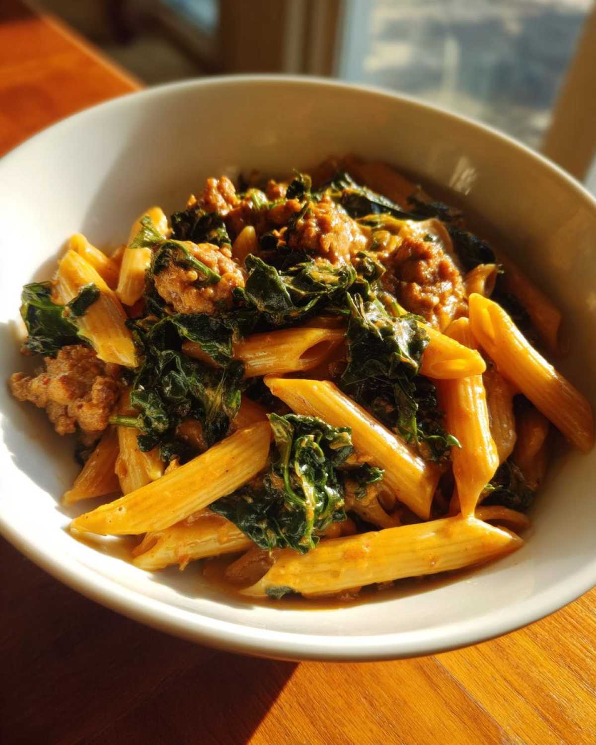 A close-up shot of a white bowl filled with One-Pot Spicy Sausage and Kale Pasta, featuring penne, crumbled sausage, and wilted kale.