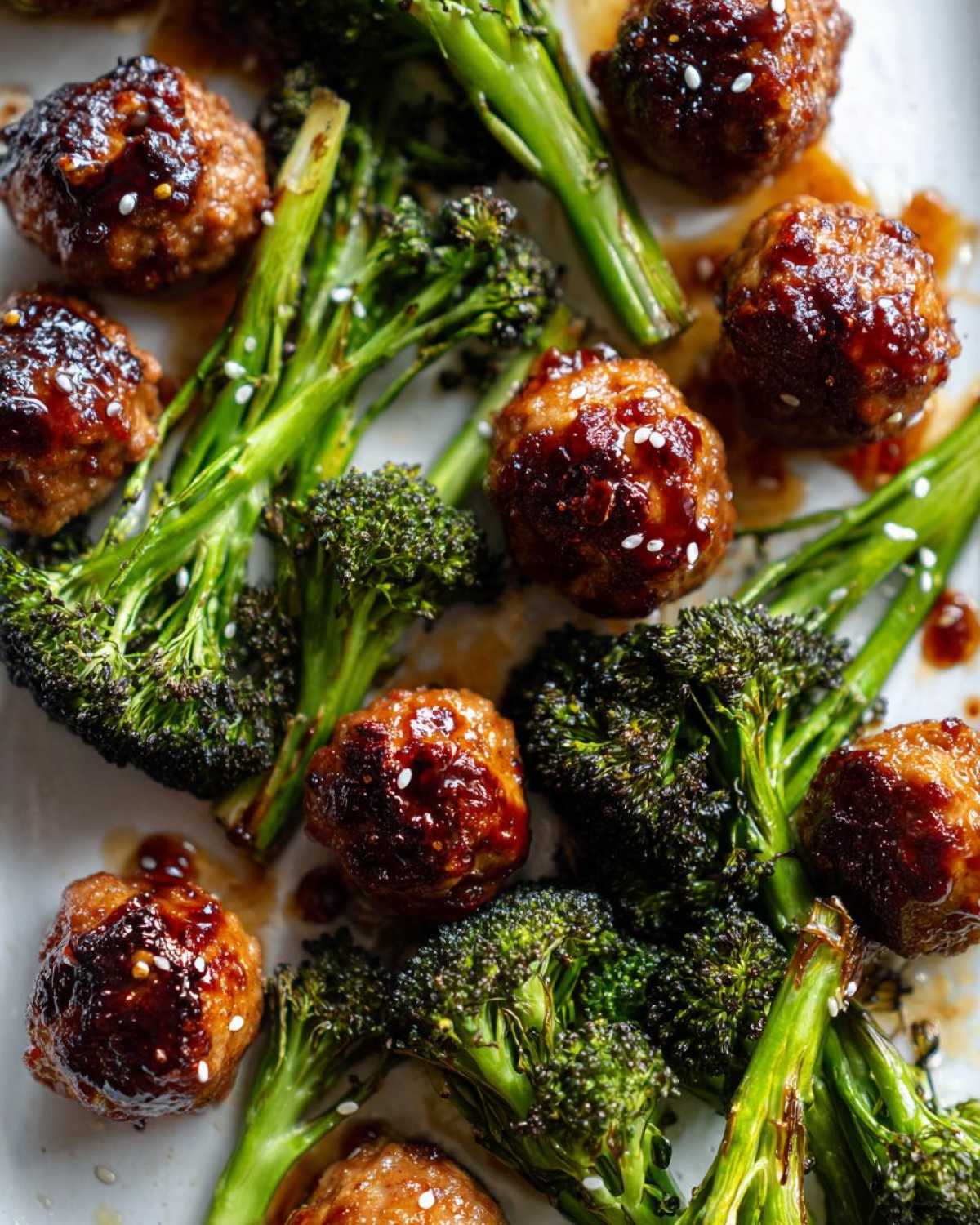 Close-up of glossy Sheet Pan Ginger Sesame Chicken Meatballs with roasted broccoli florets, sprinkled with sesame seeds.