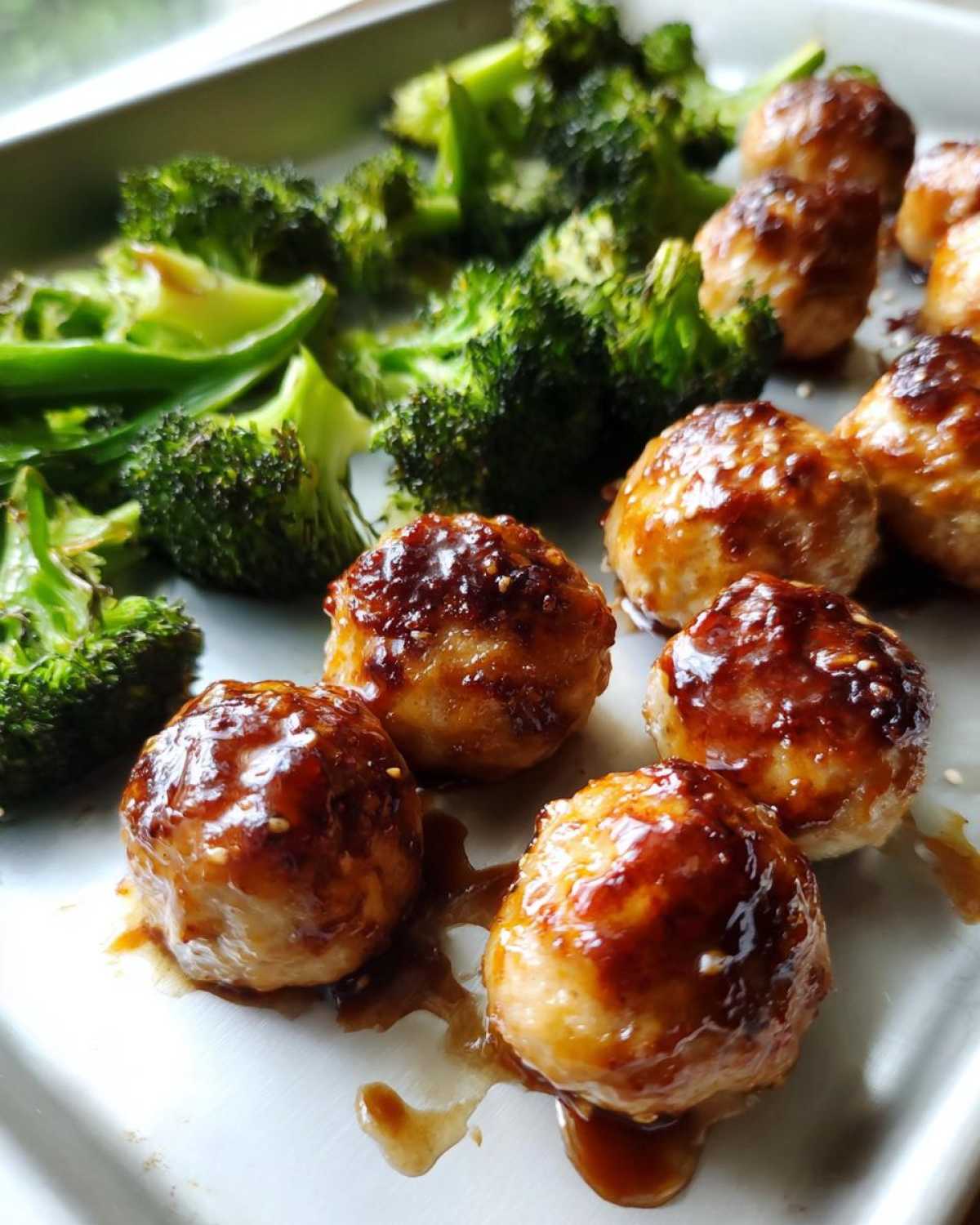Close-up of glazed Sheet Pan Ginger Sesame Chicken Meatballs with roasted broccoli florets on a baking sheet.
