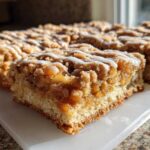 A close-up view of freshly cut apple pie bars featuring a thick layer of cinnamon apple filling, crumb topping, and a white icing drizzle.