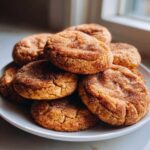 A close-up stack of freshly baked churro cookies generously coated in cinnamon sugar, resting on a white plate.