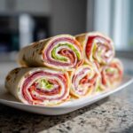 Close-up of several delicious italian pinwheels stacked on a white plate, showing layers of deli meat and lettuce.