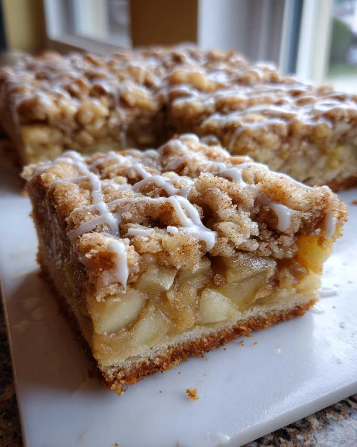 A close-up shot of a single serving of apple pie bars, showing the thick apple filling, crumb topping, and white icing drizzle.