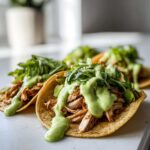 Close-up of two delicious Avocado Lime Crema Chicken Tacos topped with shredded chicken, greens, and a generous drizzle of bright green crema.