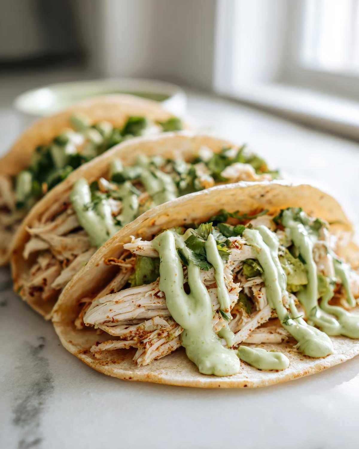 Three delicious Avocado Lime Crema Chicken Tacos lined up, topped with shredded chicken and a generous drizzle of green crema.